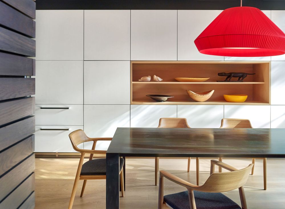 The dining room has light wooden flooring, white custom cabinetry and a bright red pendant lamp for contrast