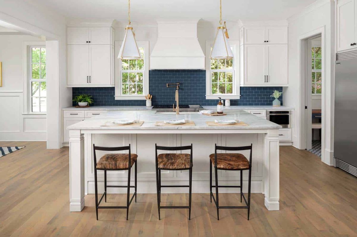 The kitchen has a large island, white walls, white furniture and a navy blue backsplash